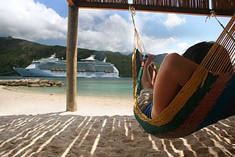 Eine Frau in einer Hängematte am Strand mit Blick auf ein vorbeifahrendes Kreuzfahrtschiff