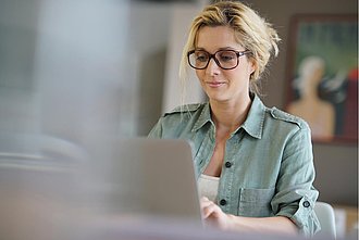 eine blonde Frau mit einer Brille arbeitet an einem Laptop